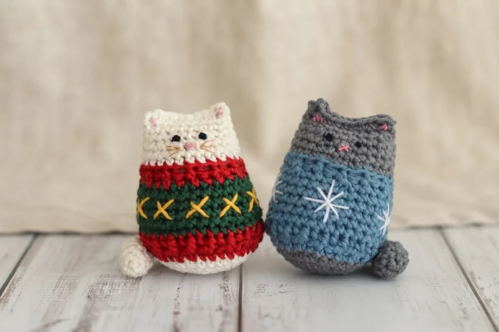 Two small crochet cats in festive sweaters on a white wooden surface, one white with red, green and yellow stitch pattern, one gray with blue sweater and white snowflake stitches