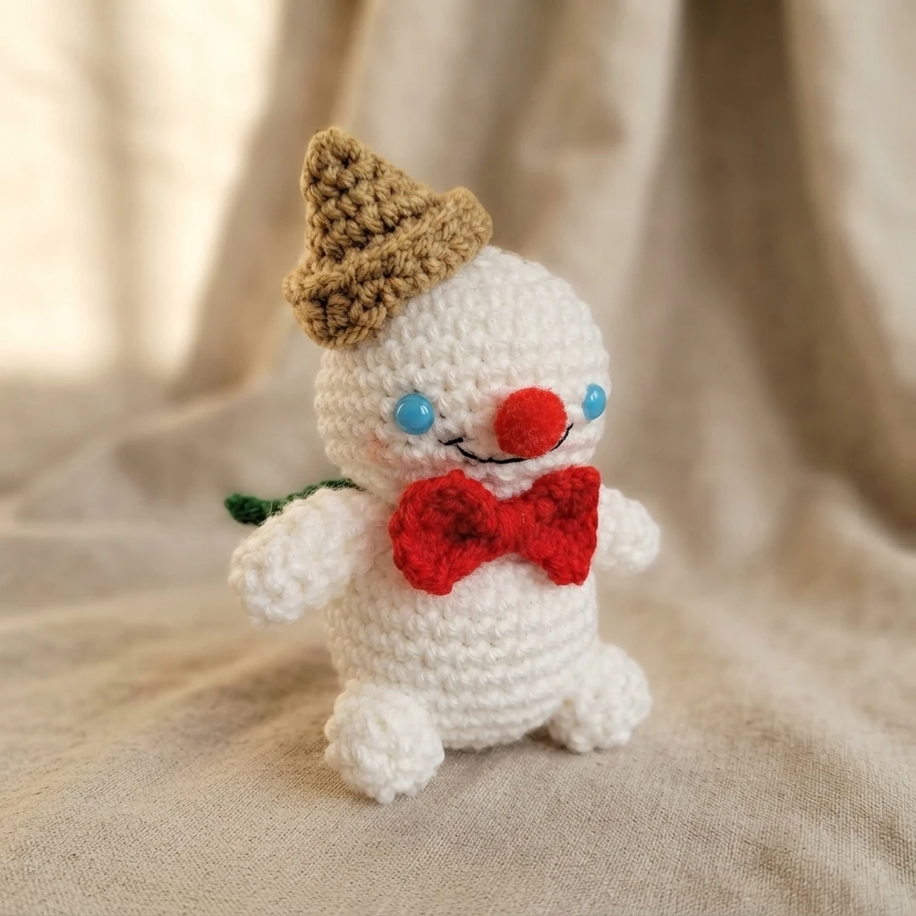 Crocheted white plush figure with a brown cone hat, blue eyes, a red nose and a red bow tie, standing on beige fabric