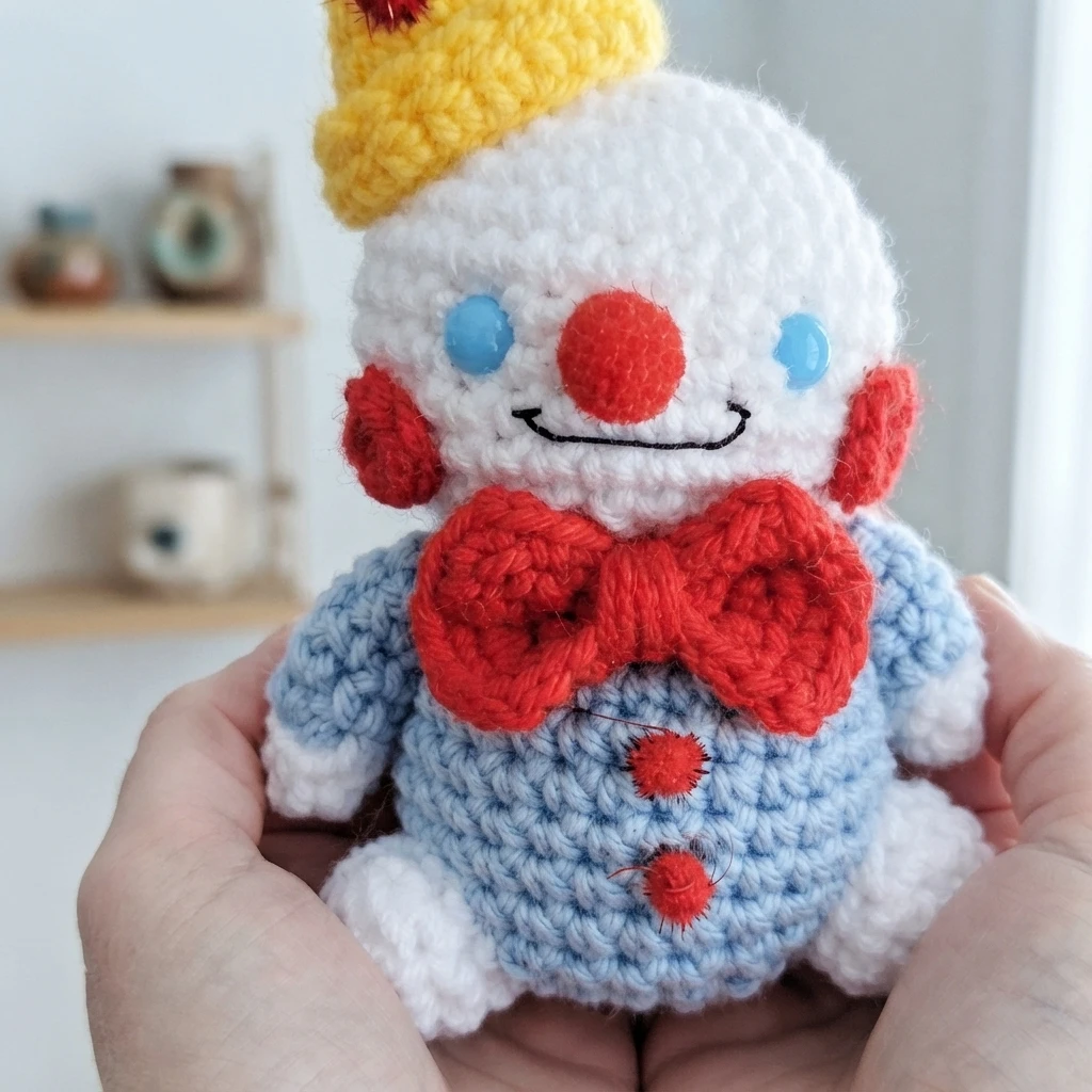 Crocheted white snowman-style plush with a yellow cone hat topped by a red pom, blue eyes, red nose and cheeks, a red bow tie and a light blue body with red buttons, held in two hands.
