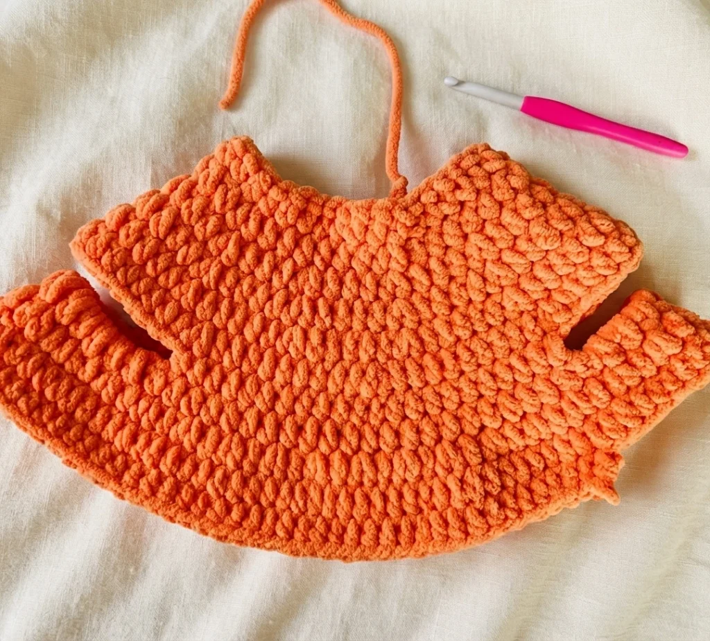 Pièce orange en cours de crochet, posée à plat avec des fils libres et un crochet rose sur un tissu beige.