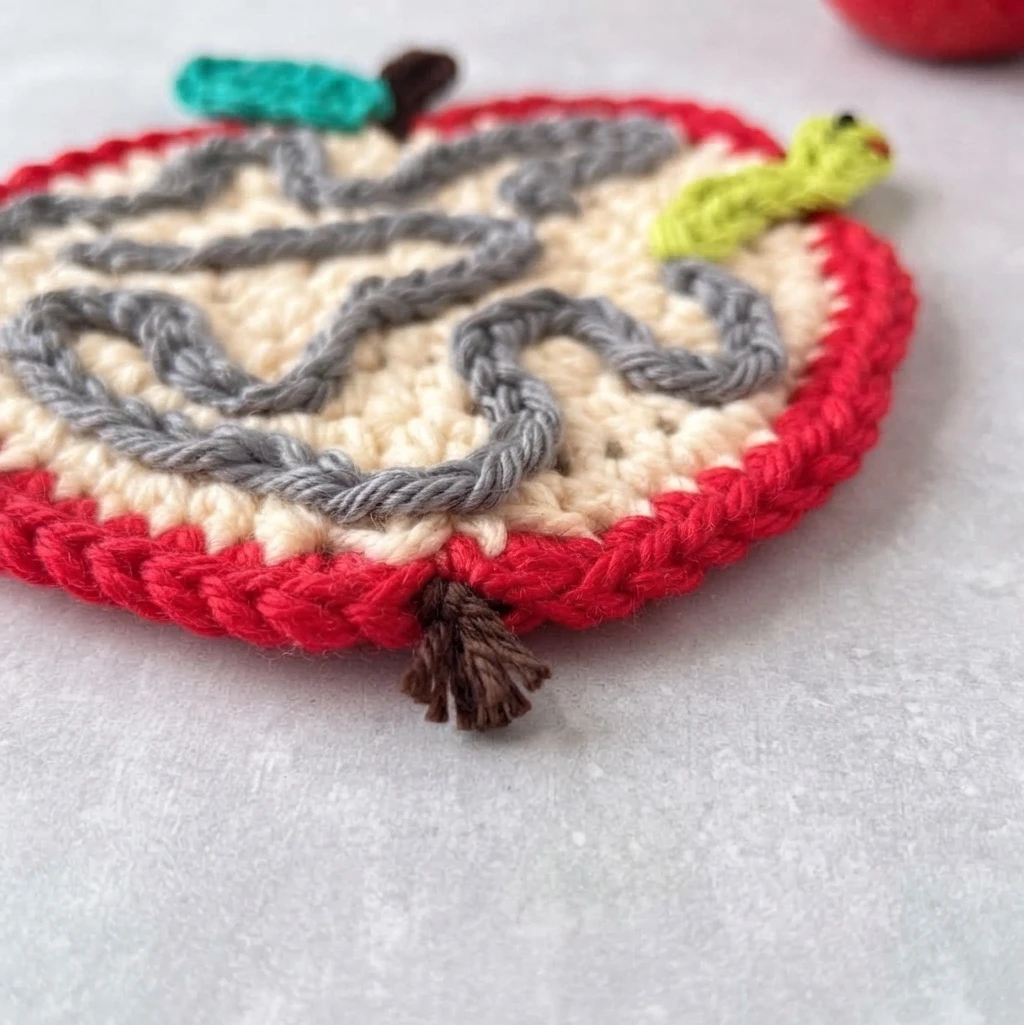 Crochet apple coaster photographed at an angle, showing a cream center with a gray yarn squiggle trail, a red border, a lime green worm, a teal leaf, a brown stem and a small brown tassel of yarn ends at the bottom blossom end.