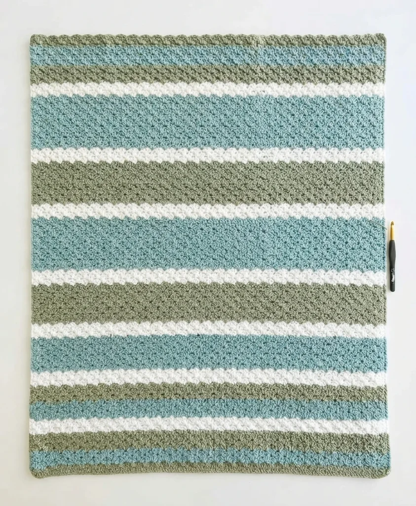 Flat lay of a striped crochet blanket in light blue, sage green and white with a crochet hook beside it