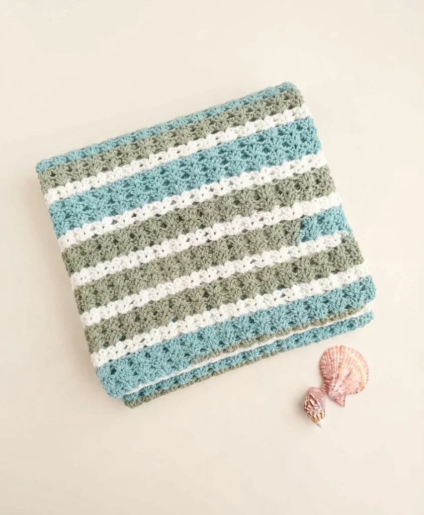 Folded striped crochet blanket in blue, sage green and white beside two seashells on a light background.