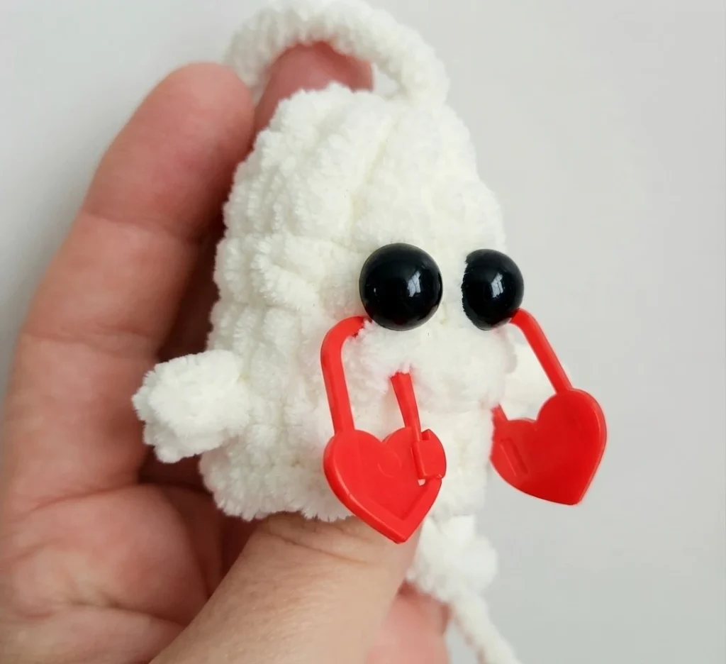 A hand holds a white crocheted ghost with black eyes and red heart-shaped stitch markers around its face.