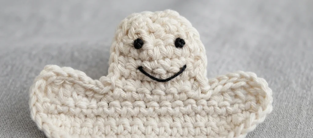 Crocheted white ghost with two black embroidered eyes and a curved black stitched smile, arms spread out to the sides, lying on a plain grey fabric background.