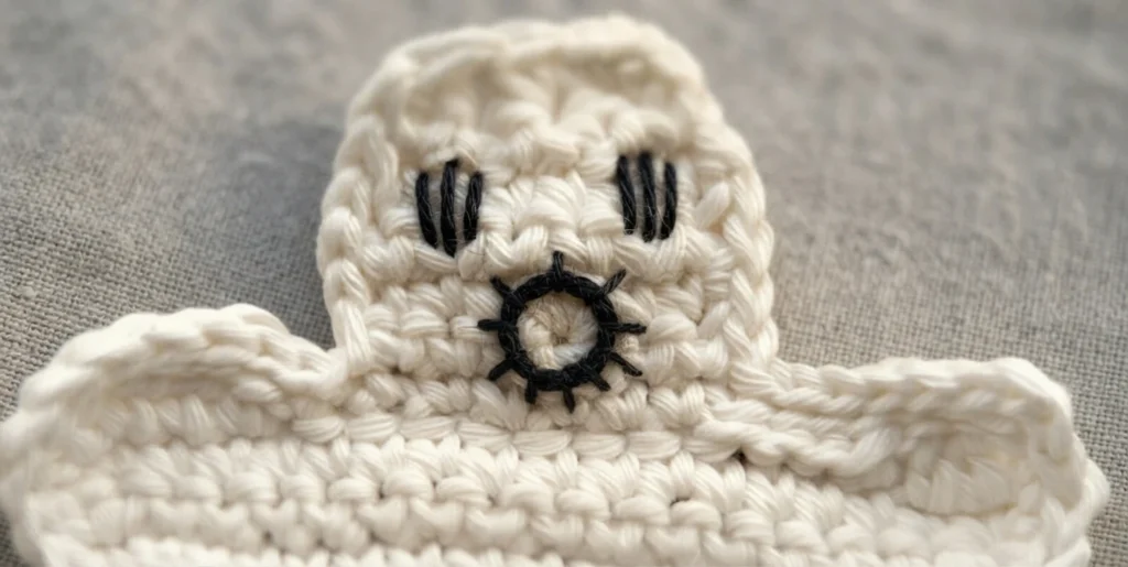 Crocheted white ghost with black stitched eyes made of short vertical lines and an open round mouth outlined in black thread, resting on a grey woven fabric surface.