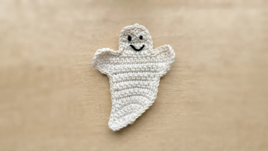 White crocheted ghost applique with two small black eyes and a black stitched smile, lying flat on a light wood surface.