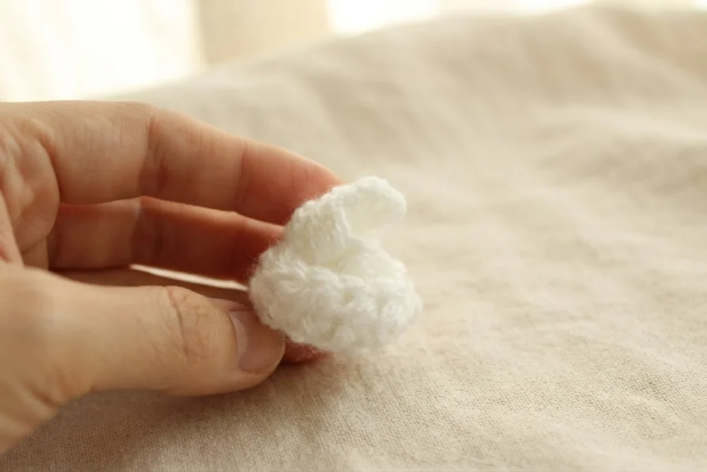 A hand holding a small white crocheted whipped cream swirl with a curled tip, photographed against a soft beige linen background.