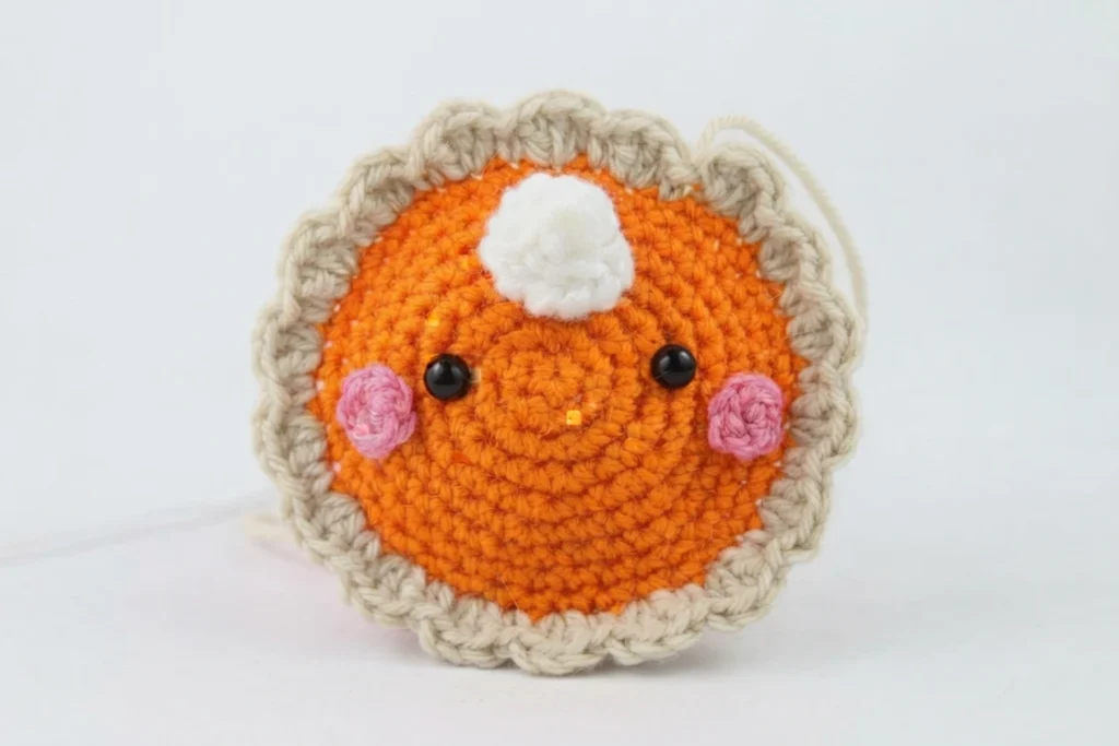 Crocheted amigurumi pumpkin pie with an orange face, a tan scalloped crust edge, two pink cheeks, two black bead eyes and a white whipped cream swirl, photographed from the front on a white background