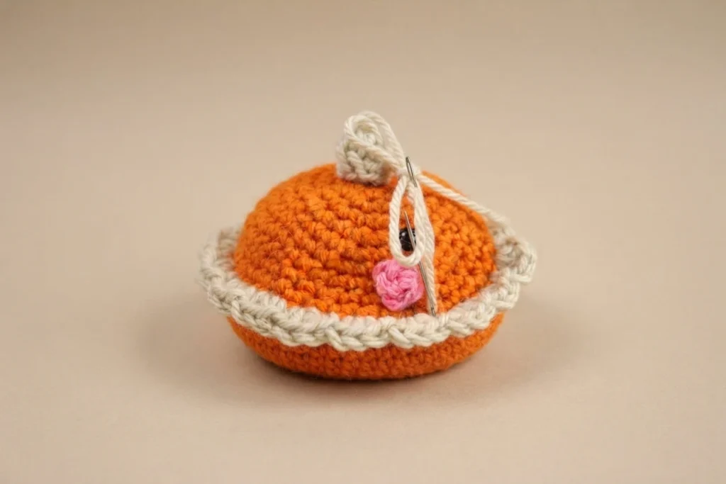 Crocheted pumpkin pie amigurumi with an orange top, a tan crust rim, a white whipped cream swirl, one black safety eye, a pink cheek and a tapestry needle resting against it.