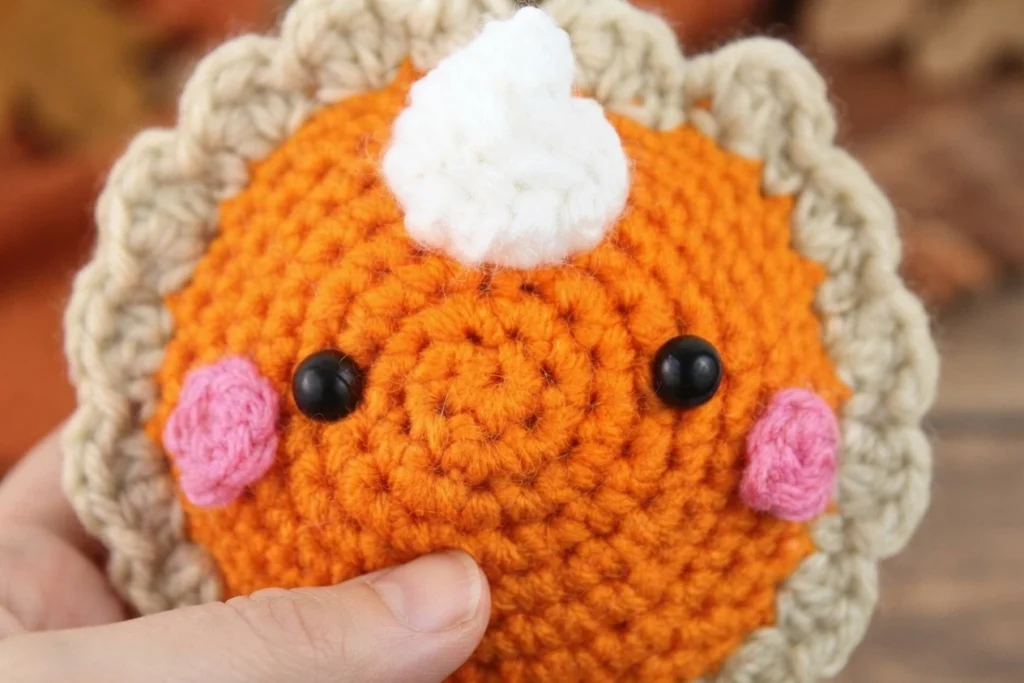 Hand holding a crocheted pumpkin pie amigurumi with an orange face, black safety eyes, pink cheeks, a white whipped cream swirl on top and a tan scalloped crust.