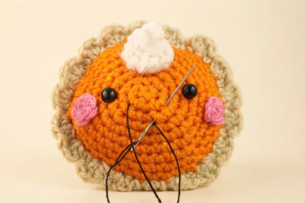 Crochet pumpkin pie amigurumi with orange filling, tan scalloped crust, pink cheeks, black safety eyes, white whipped cream on top, and a needle threaded with black yarn stitching the smile.