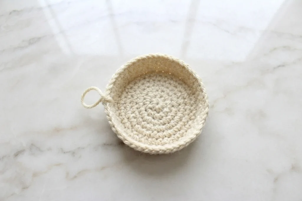 Small crocheted tan bowl shape with a spiral base and a yarn loop at the rim, photographed from above on a light surface.
