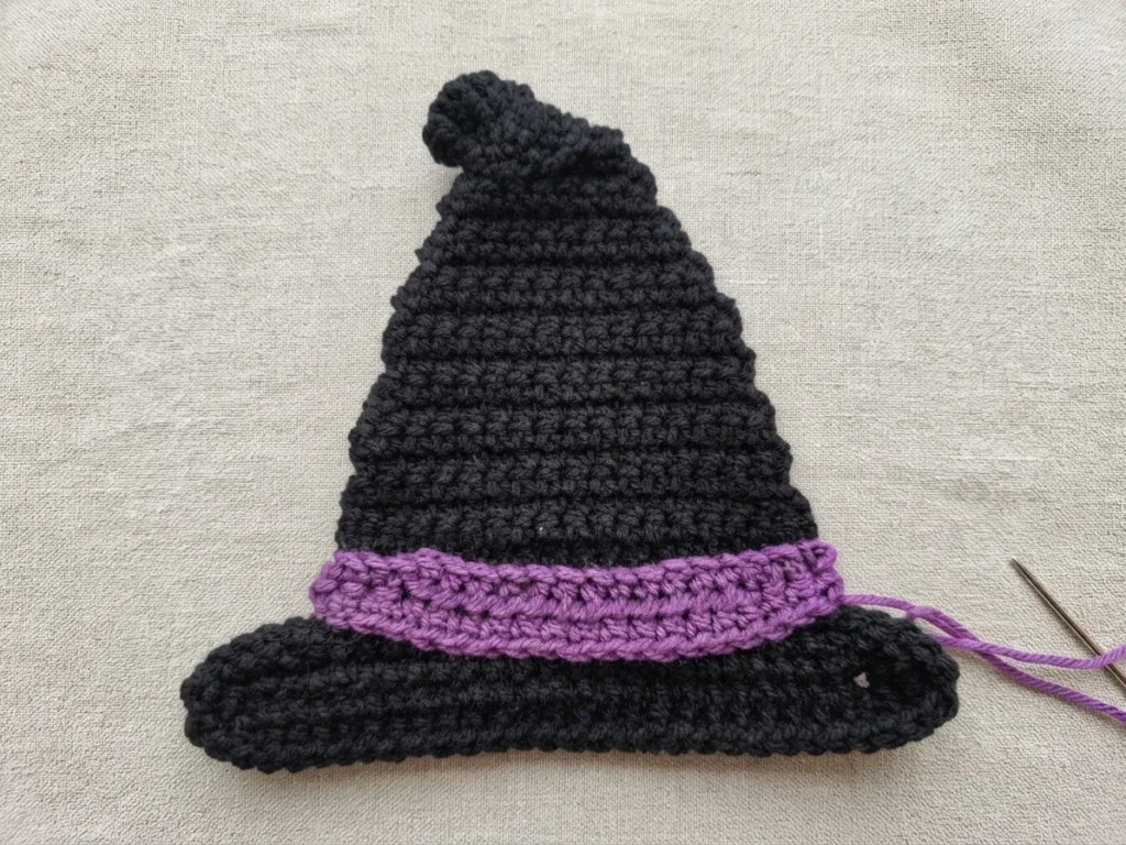 Black crocheted witch hat with a purple band around the brim, resting on a light textured fabric, with a tapestry needle and a strand of purple yarn at the lower right.