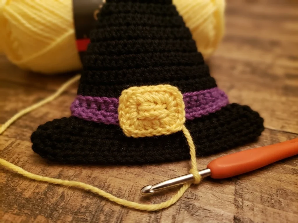 Crocheted black witch hat with a purple band and a yellow crocheted buckle, resting on a wooden surface beside yellow yarn and an orange crochet hook.