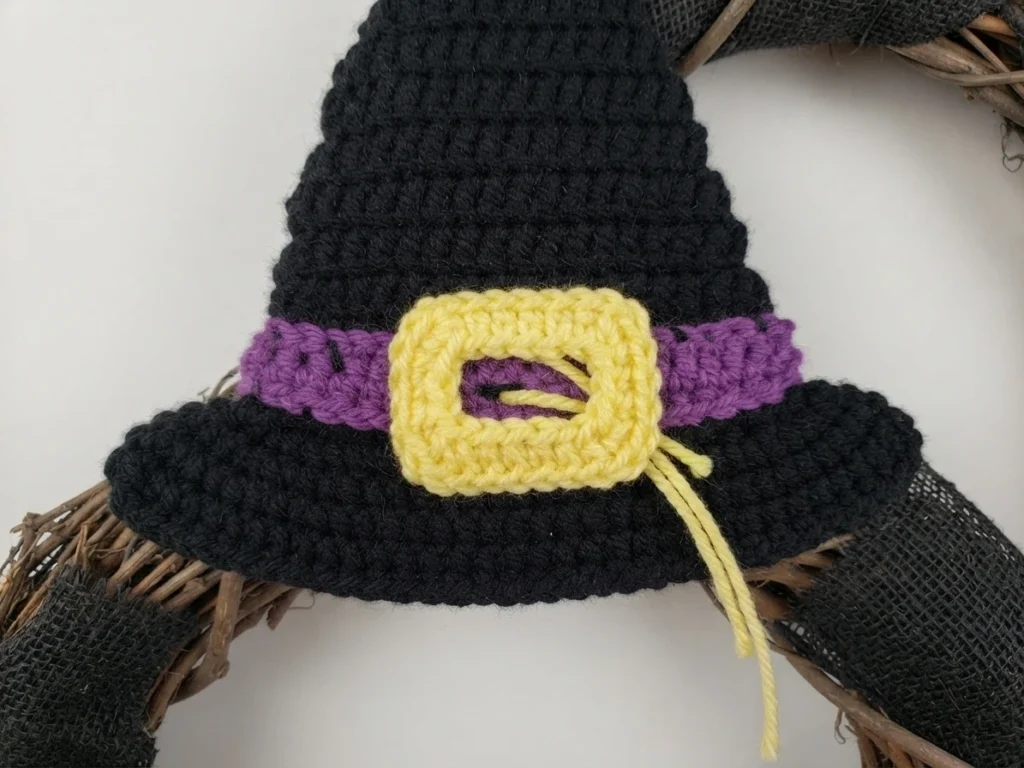 A yellow crocheted rectangular buckle sewn onto the purple band of a black crocheted witch hat, with long yellow tails hanging, resting on a wreath form covered with black mesh.