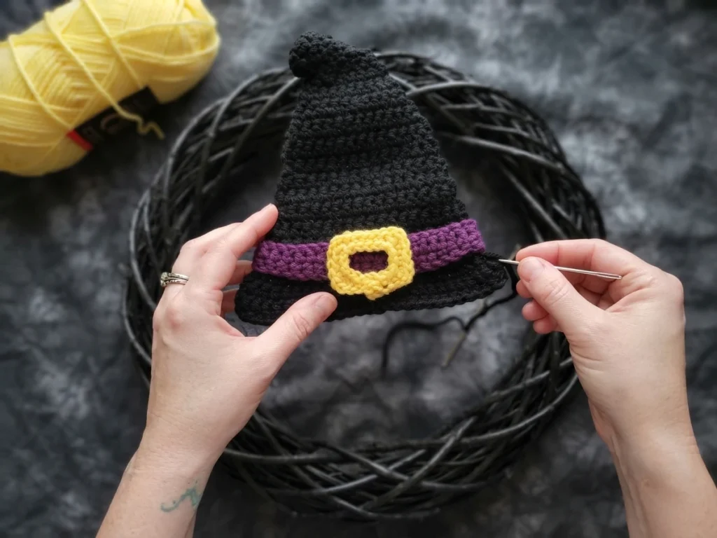 Two hands holding a crocheted black witch hat with a purple band and a yellow buckle, using a tapestry needle to stitch the buckle, with a ball of yellow yarn at the left and a dark textured background.