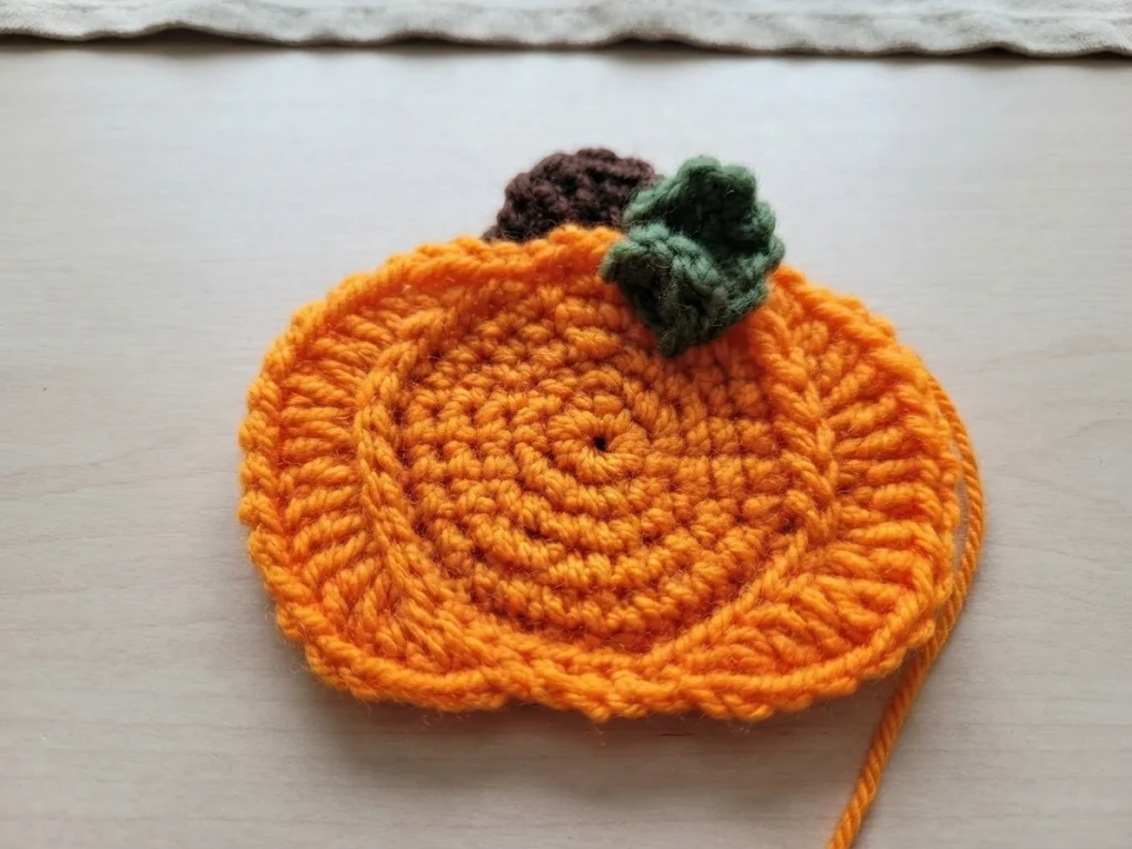 Crocheted orange pumpkin motif with a brown stem and a small green curled leaf, worked in a spiral of stitches, resting on a light wood surface with an orange yarn tail at the lower right.