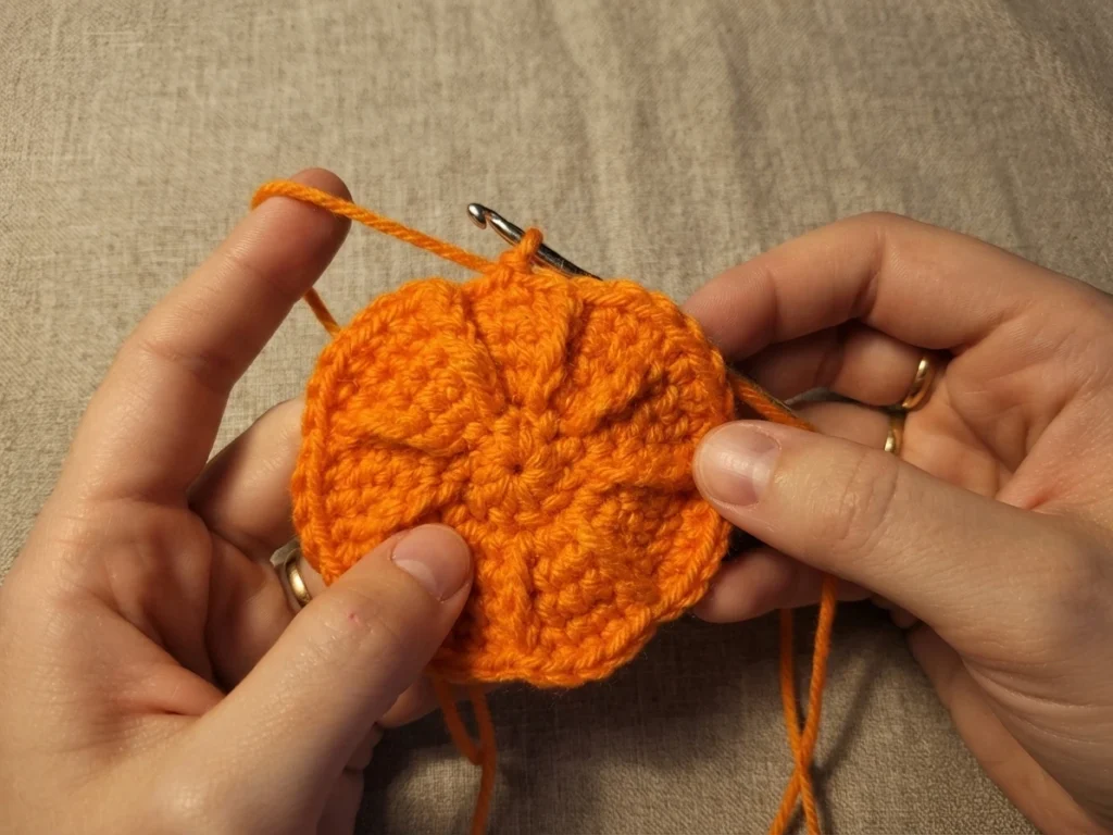 Two hands holding a small round orange crocheted piece with a metal crochet hook, working the pumpkin shape in progress against a beige fabric background