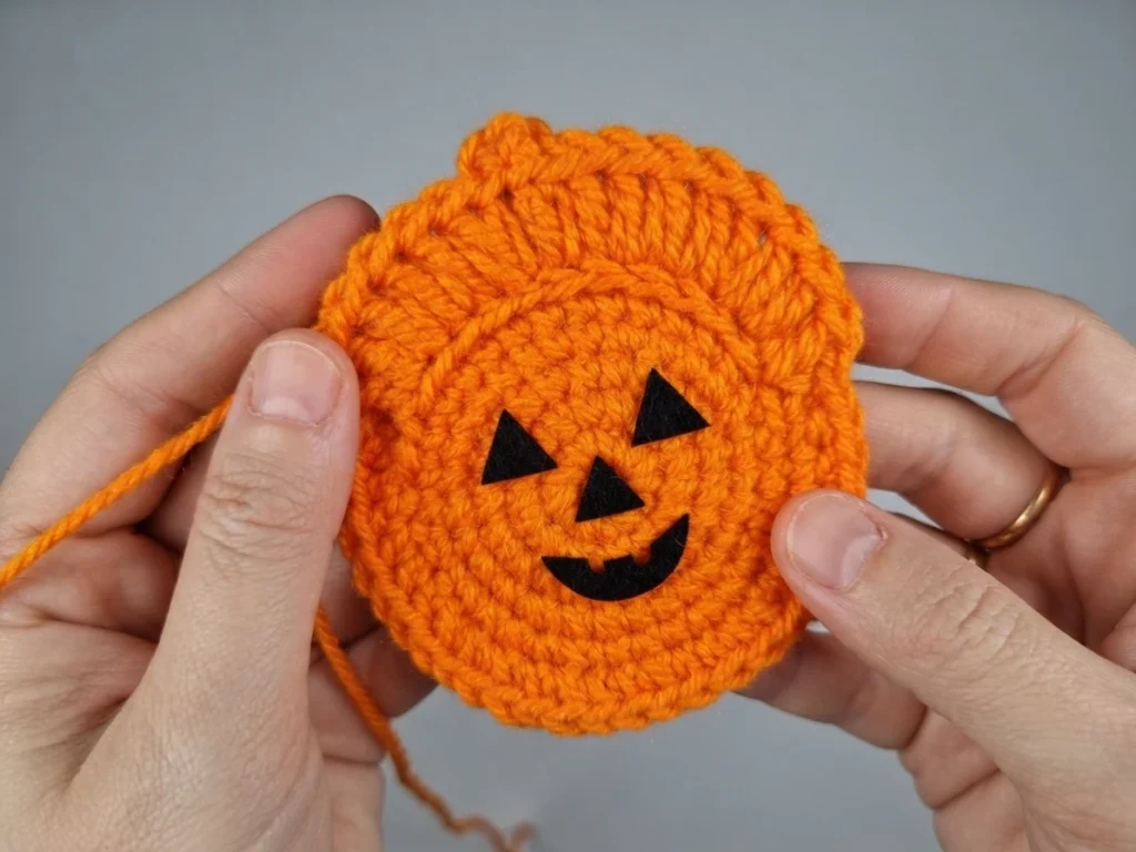 Hands holding an orange crocheted pumpkin circle with a black felt jack-o-lantern face, shown against a plain grey background with a loose yarn end at the left.
