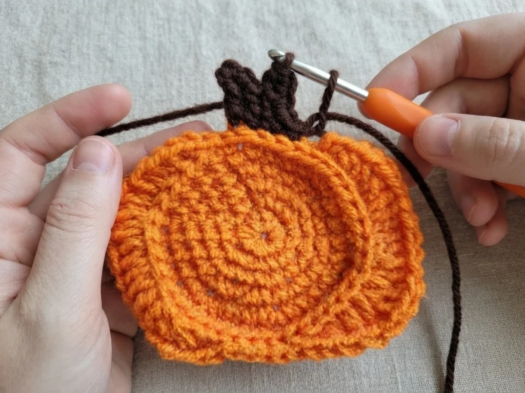 Hands holding a crocheted orange pumpkin while a dark brown stem is worked with an orange-handled crochet hook