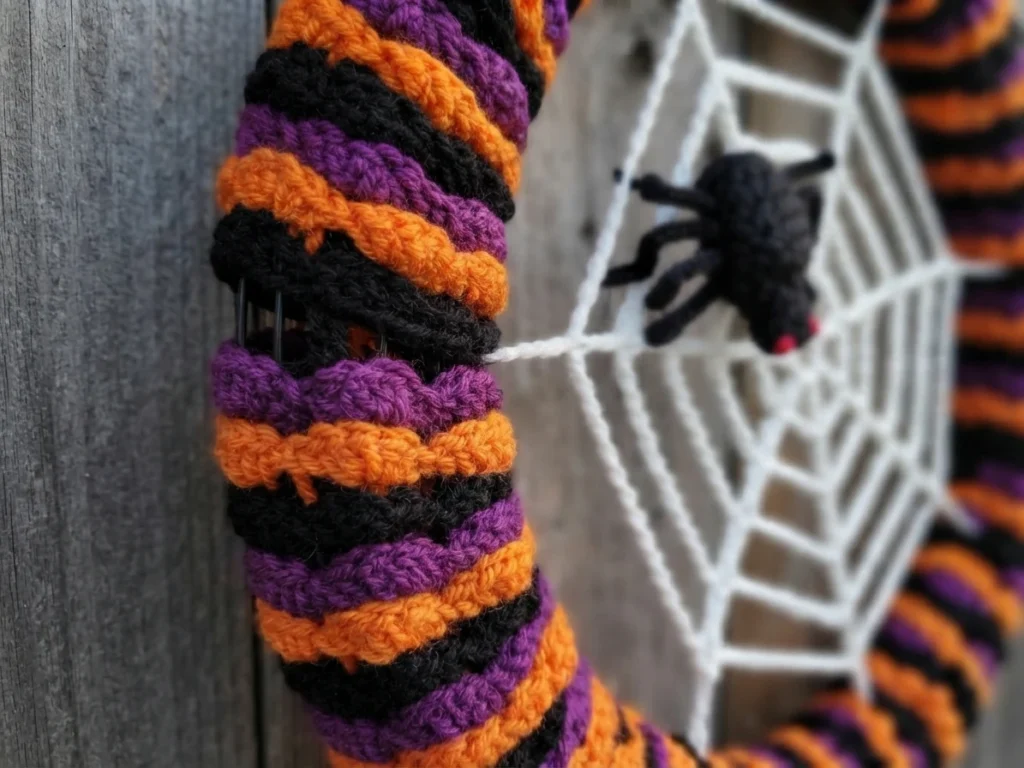 Close-up of a crocheted Halloween wreath showing spiralling black, purple and orange shell stripes wrapped around a wire frame, with a white crocheted spiderweb and a black crocheted spider visible in the blurred background.