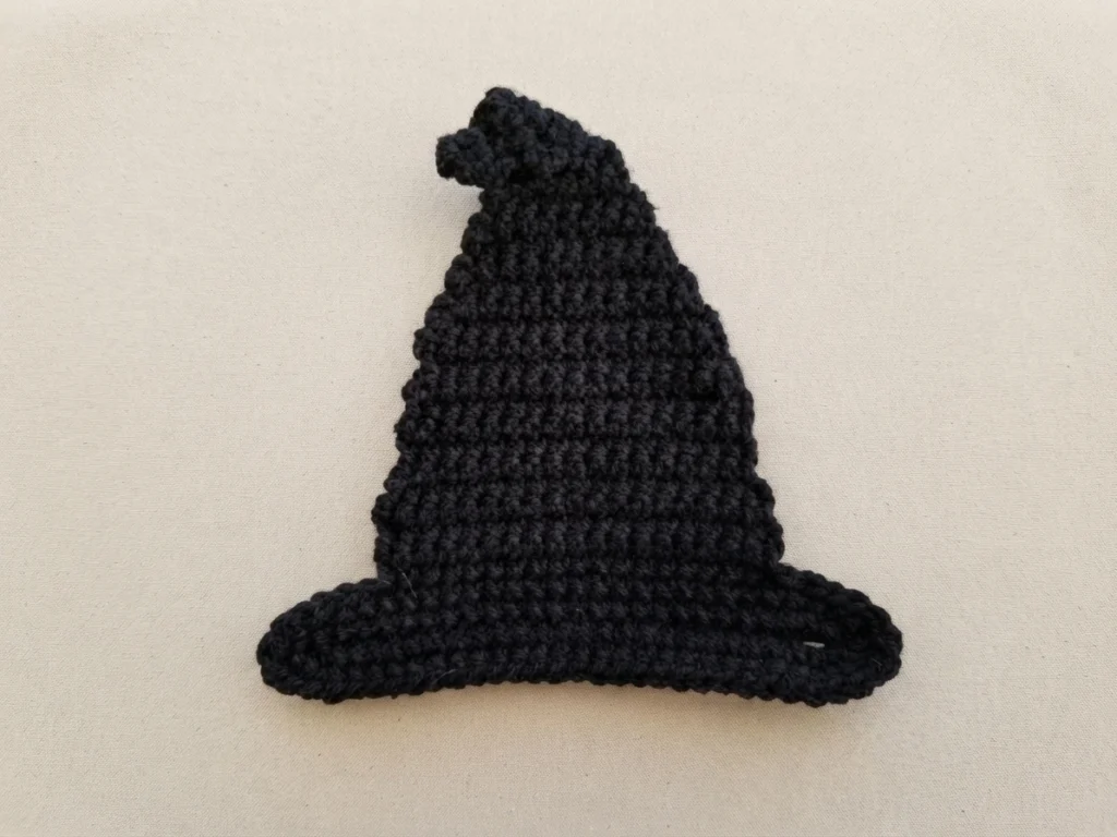Black crocheted witch hat with a curled tip and a wide brim, lying flat on a plain cream fabric background.
