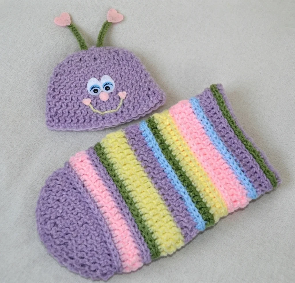 Crocheted lavender love bug newborn photo prop set: bug hat with googly eyes, stitched smile and heart-tipped green antennae, laid beside a pastel-striped crocheted cocoon.