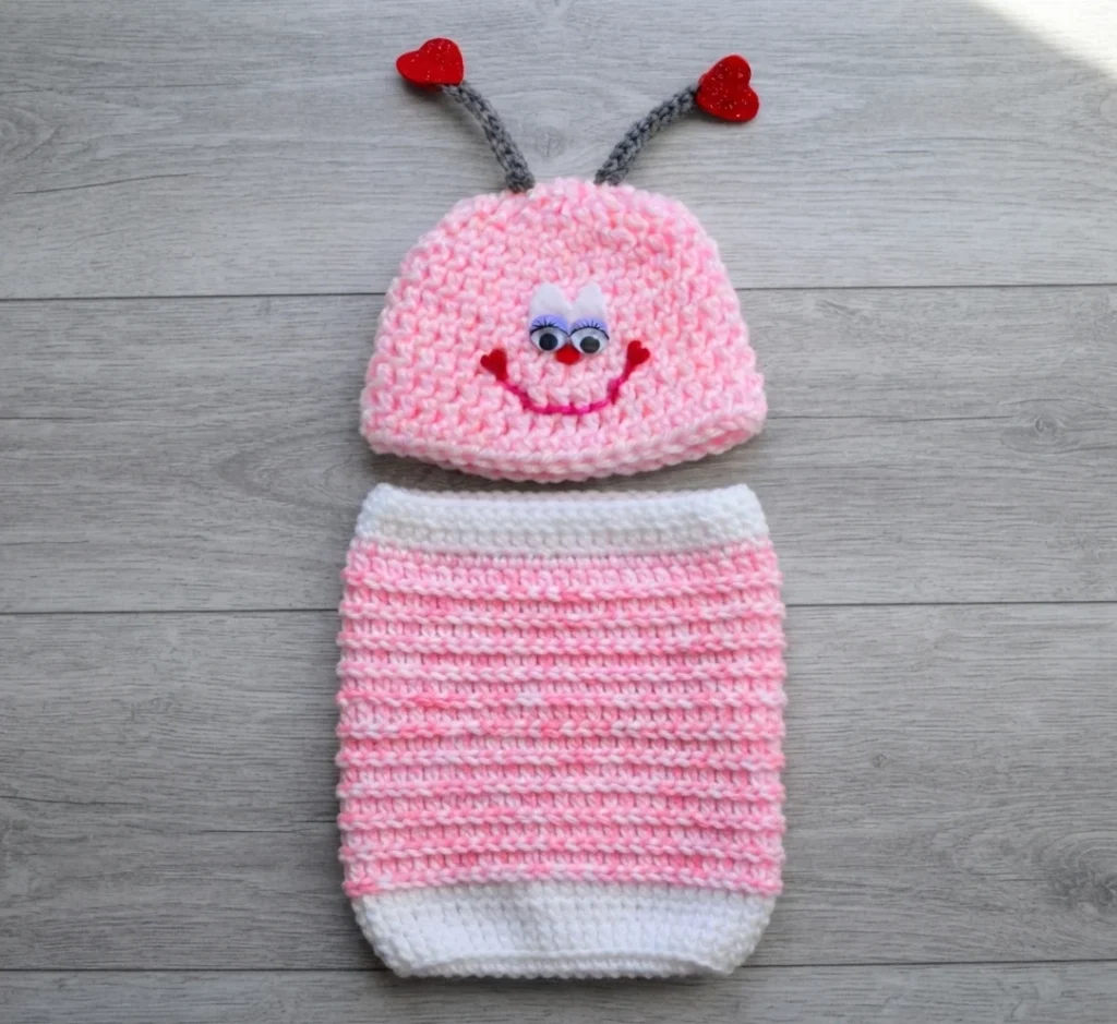 Pink crochet love bug hat with grey antennae topped with red hearts, googly eyes and a smiling face, placed above a pink and white striped crochet cocoon on a grey wooden surface.