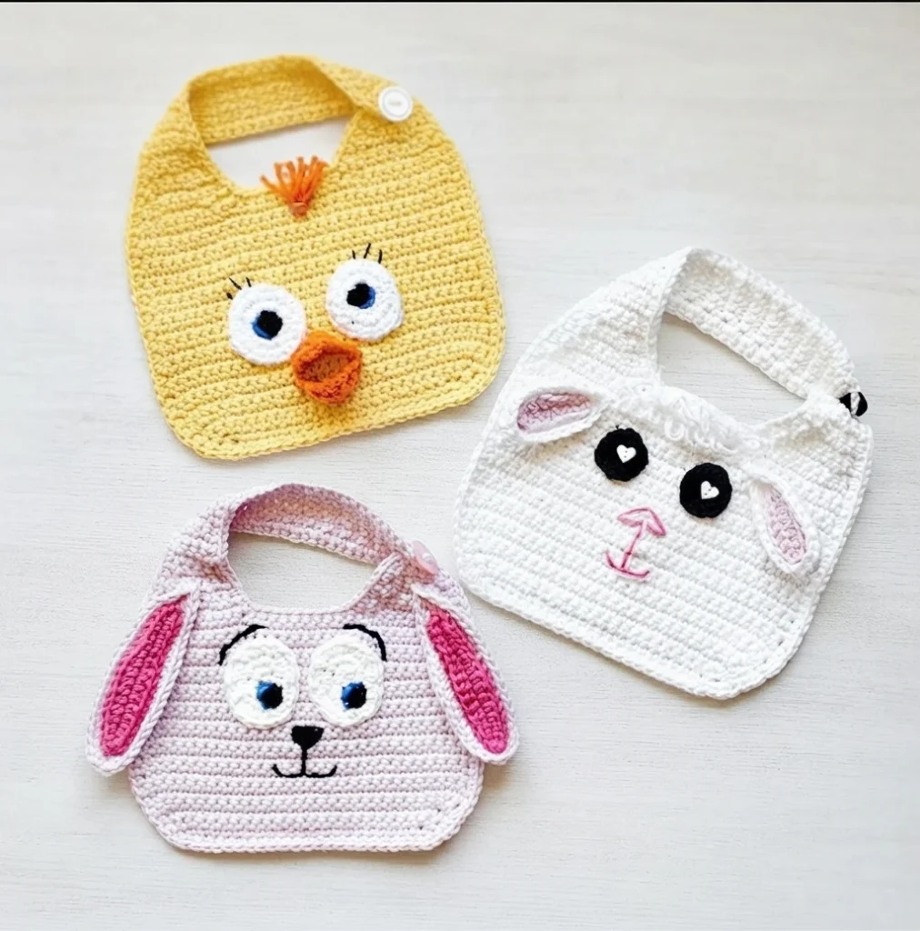 Three crocheted baby bibs with animal faces: a yellow chick, a white lamb, and a pink bunny, each with a button strap.