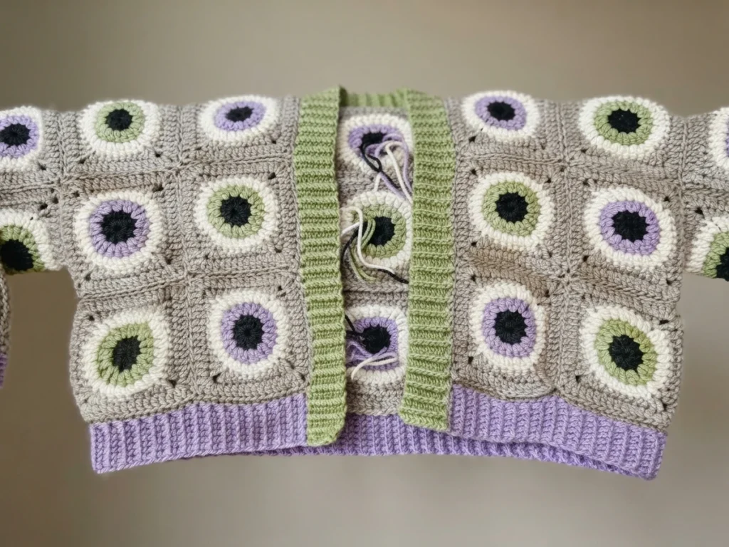 Lavender, green and grey granny square cardigan with eyeball motifs laid flat, showing a green collar band, purple ribbed hem and loose yarn tails at the center.