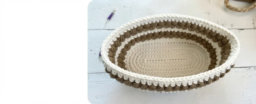 Top view of an unfinished oval crochet bag with white and brown puff stitch stripes on a white wooden surface