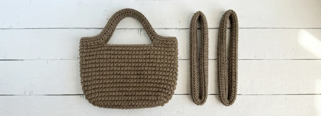 Crocheted brown tote bag body with one attached handle and two separate finished tube handles lying on a white wooden surface.