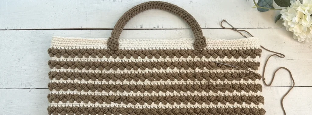 Striped braided crochet tote bag laid flat with its handle attached to the top border, a yarn needle and loose thread resting beside it