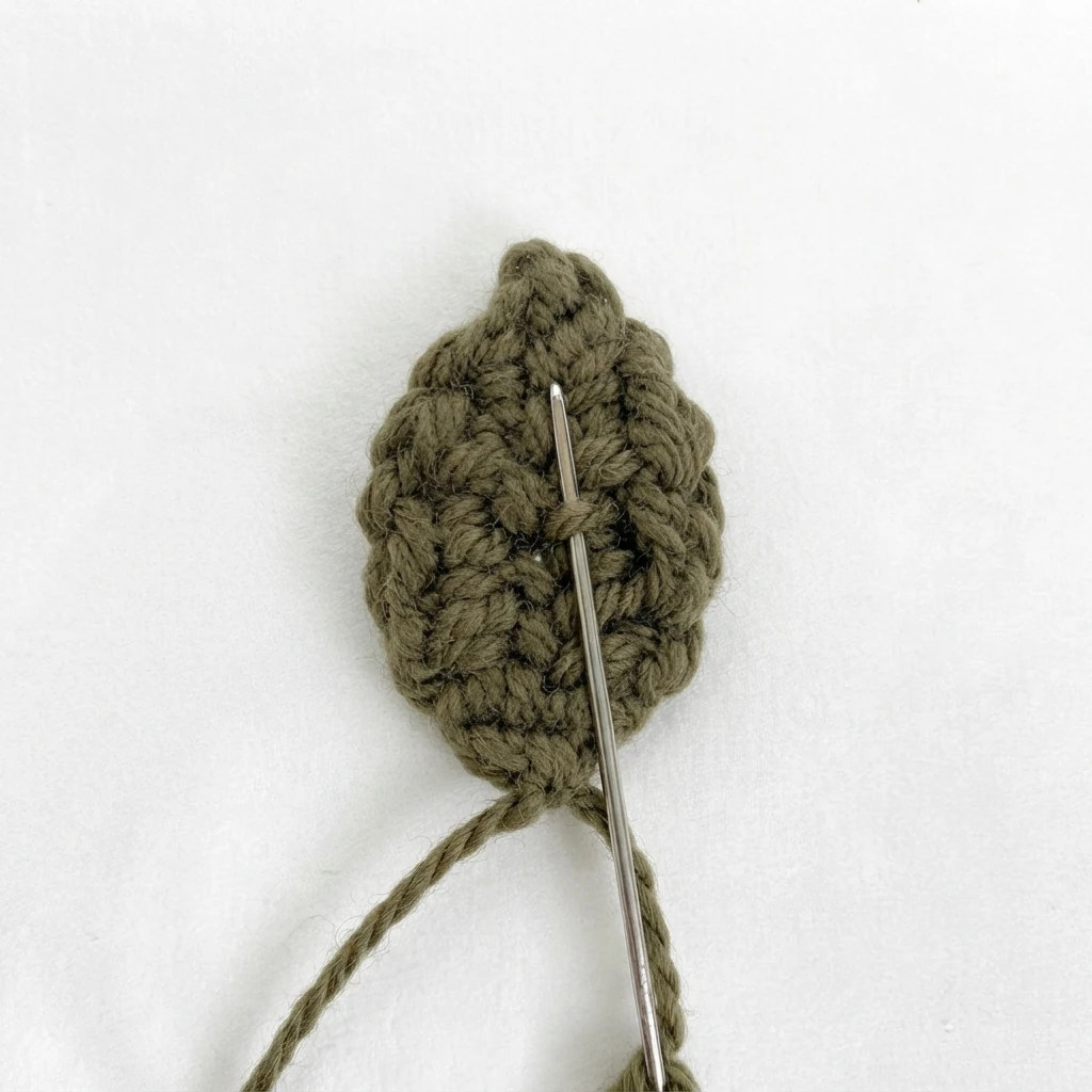 Olive green crocheted leaf with a metal tapestry needle and yarn tails on a white background.