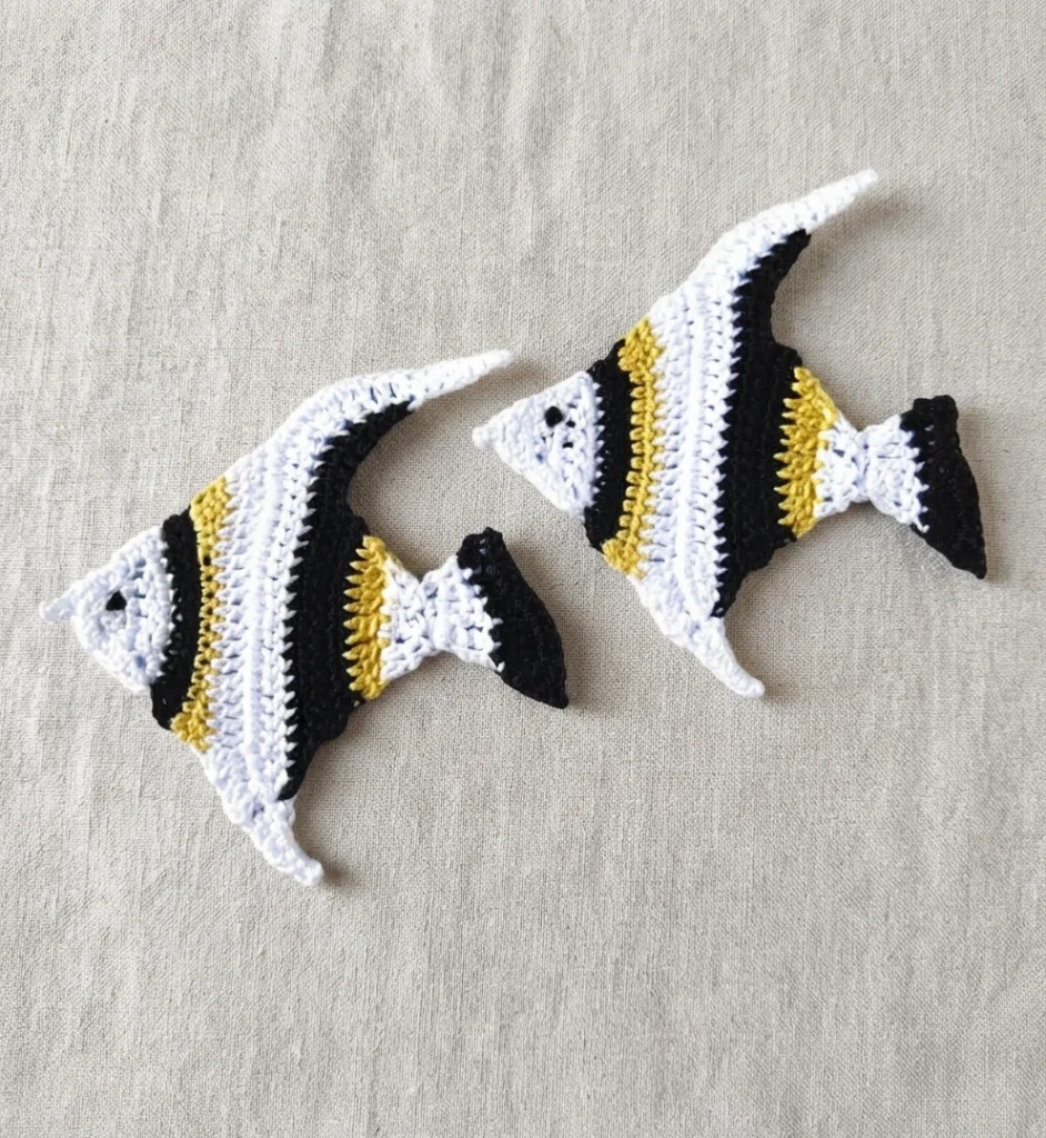 Two crochet tropical fish appliques in white, black and yellow on a beige fabric background.