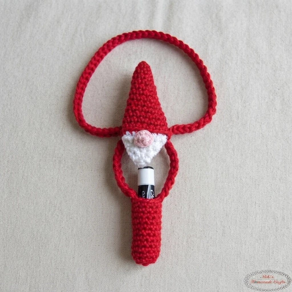 Finished crocheted Santa gnome lip balm holder in red with a pointed hat, white beard, pink nose and a long red cord loop, with a lip balm tube tucked inside the red holder