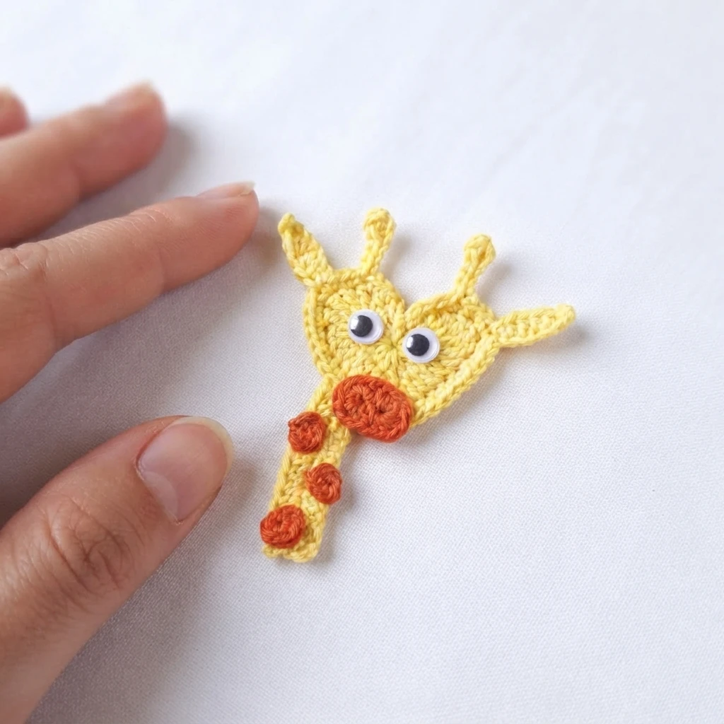 Yellow crochet giraffe applique with orange muzzle and spots lying on white fabric beside fingertips.