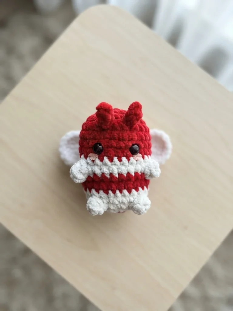 Single red crochet bee amigurumi with a white candy cane striped band around its middle, small red antennae, white wings, black safety eyes and pink cheeks, sitting on a light wooden surface indoors.