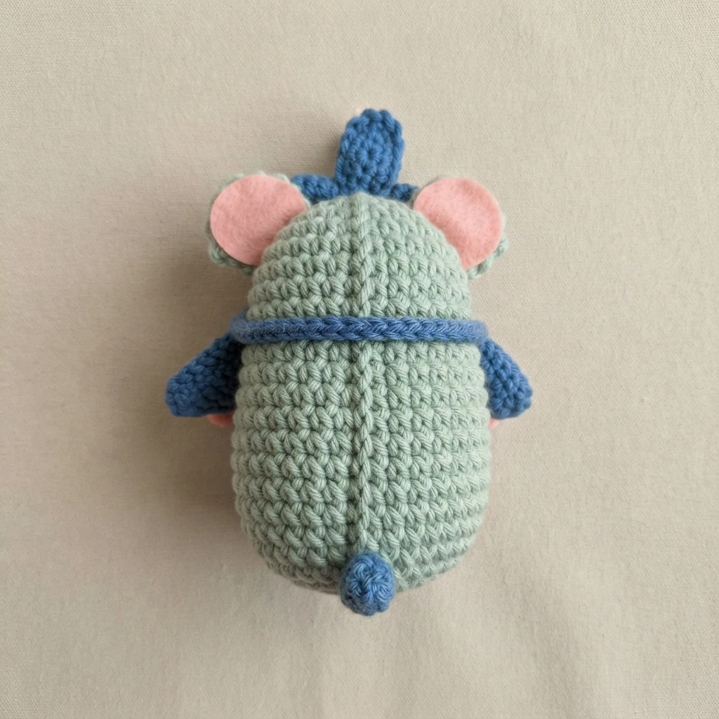 Crocheted mint green amigurumi photographed from above on a plain light background, with blue crocheted ears, blue side limbs, a blue band around the middle and a small blue topknot, and pink felt inner ears.