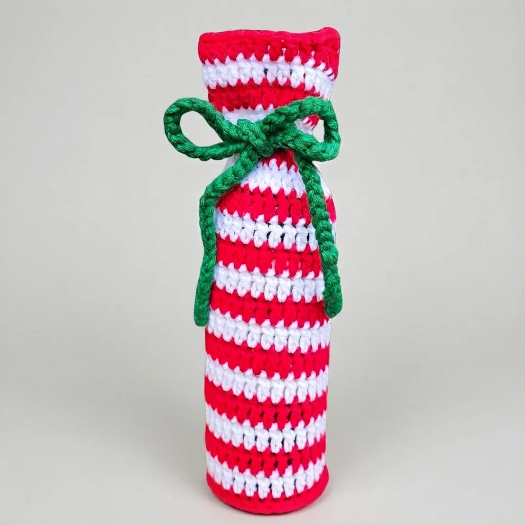 Crocheted wine bottle gift bag in red and white candy cane stripes, gathered at the neck with a green yarn bow and trailing ties, standing upright on a plain light gray background.