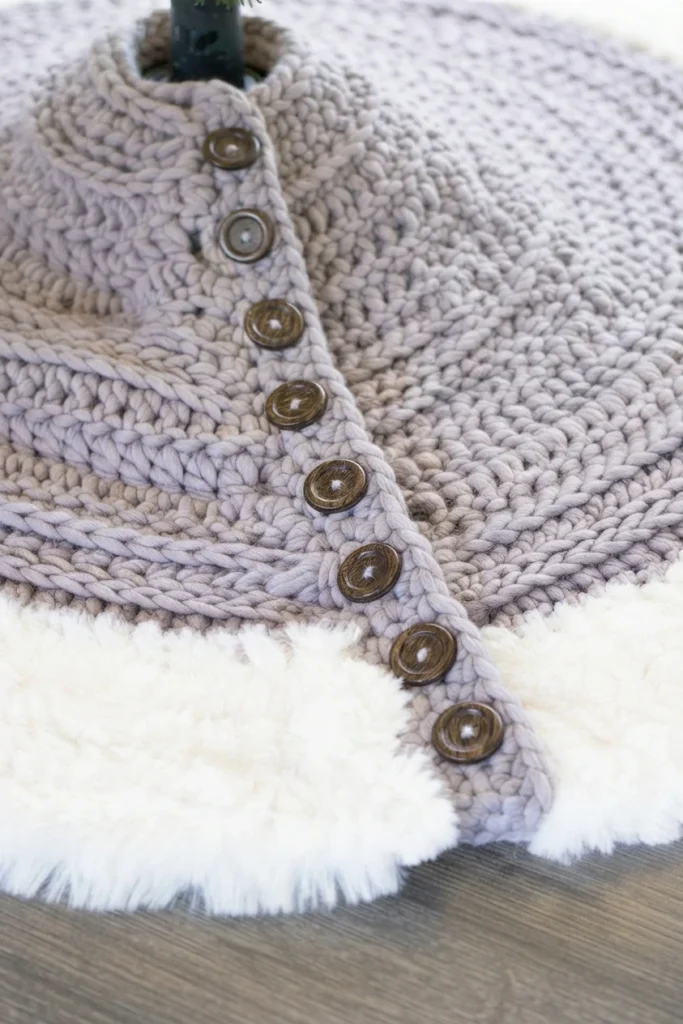 Close-up of a chunky gray-mauve crocheted tree skirt with white faux-fur trim and wooden buttons around a Christmas tree base.