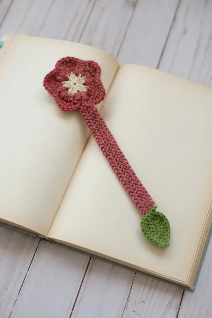 Crocheted hibiscus bookmark with a dusty rose flower with a cream center, a long textured pink strip, and a small green leaf at the end, lying on an open book with blank pages.