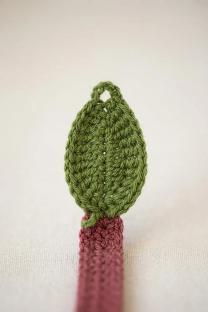 Close-up of a small crocheted green leaf joined to the tip of a mauve crocheted bookmark strip, on a plain off-white background.