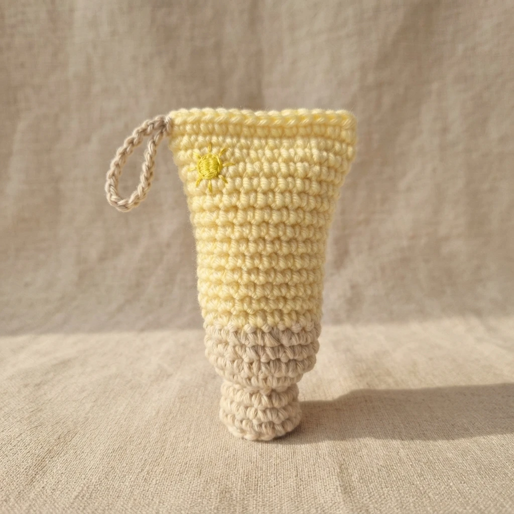Crochet sunscreen bottle in pale yellow and beige with a small embroidered sun and a hanging loop, standing on beige fabric.