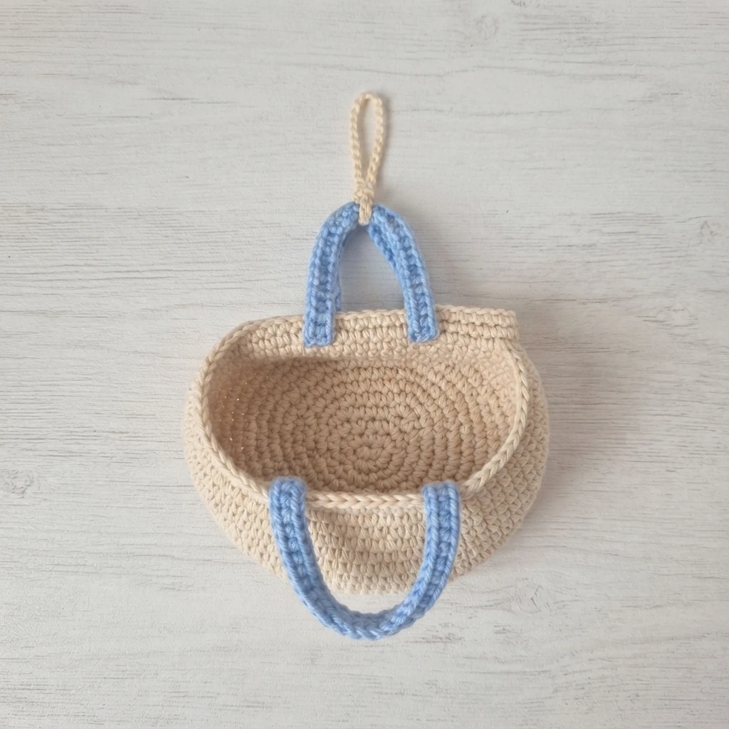 Small beige crocheted tote bag with light blue handles and a beige hanging loop, photographed from above on a whitewashed wooden surface