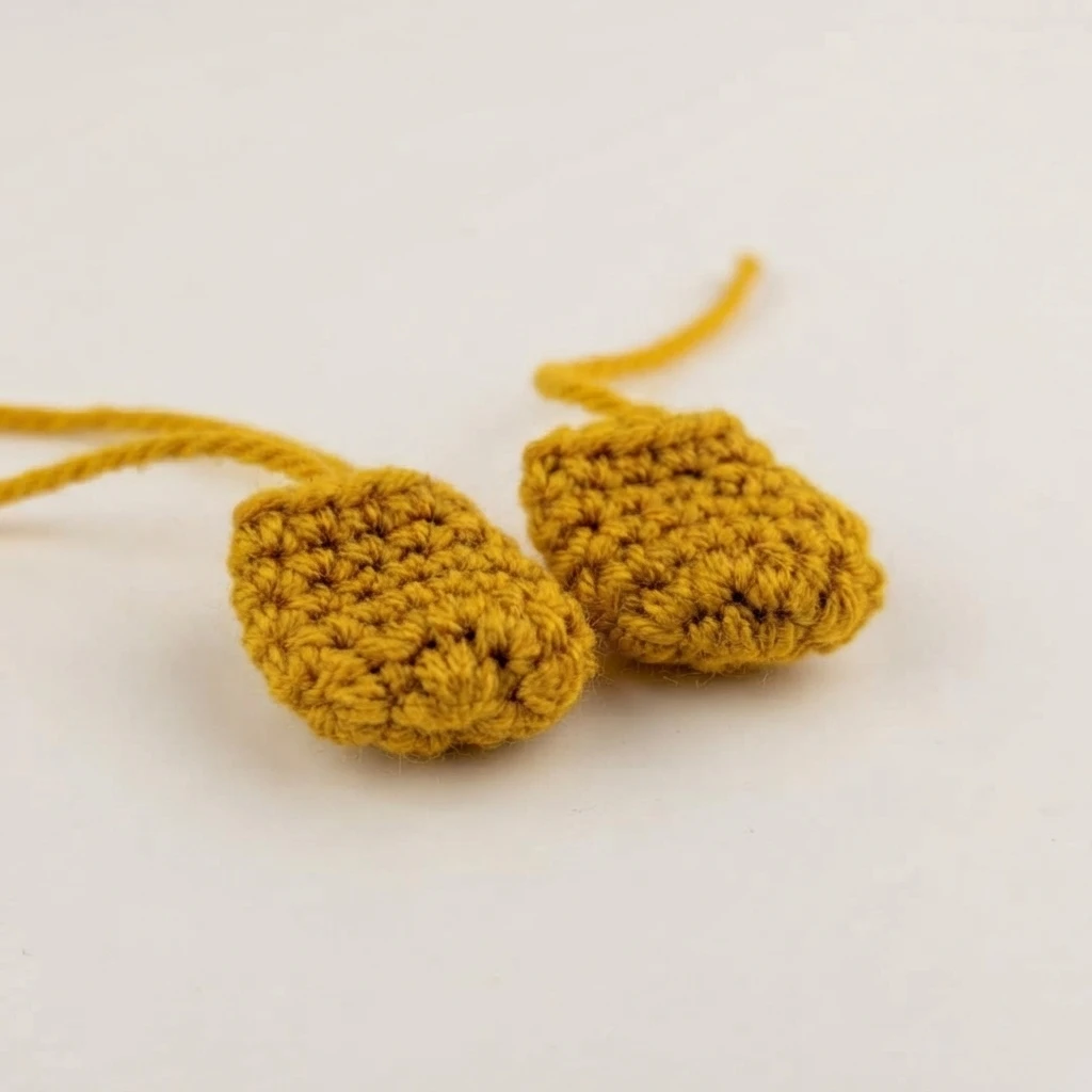 Two small mustard yellow crocheted foot pieces with long yarn tails, lying side by side on a plain white surface.