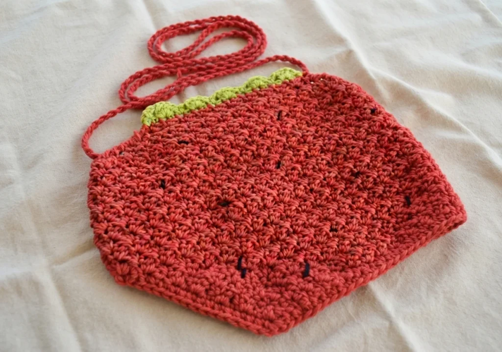 Red crocheted strawberry-shaped halter top with green trim, scattered black seed stitches and long red straps, shown lying flat on fabric.