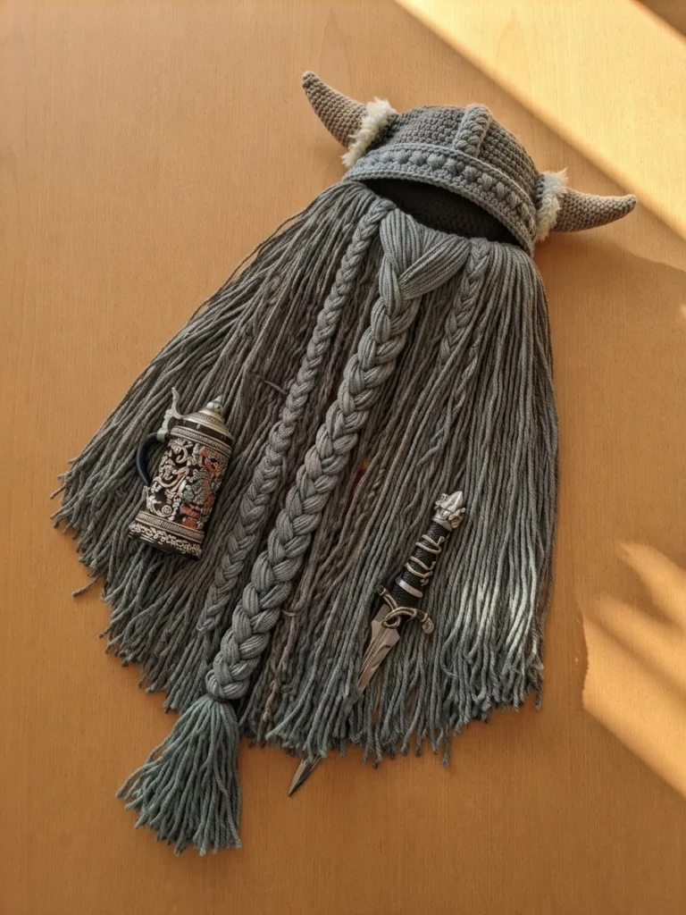 Grey crocheted Viking helmet with horns and long grey braided beard laid flat on a wooden surface beside a decorated stein and a dagger.
