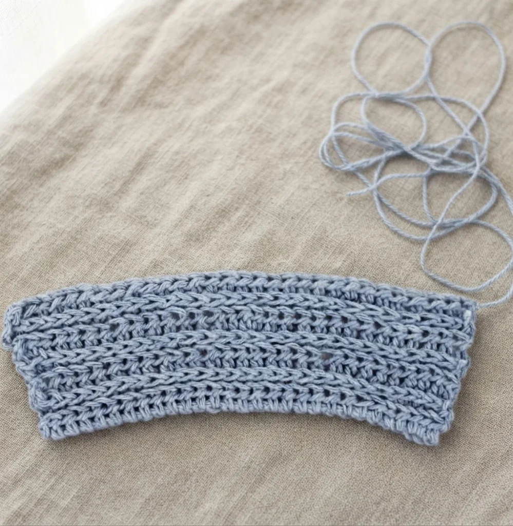 Flat blue crocheted coffee sleeve panel worked in rows, with a loose strand of matching yarn coiled in loops at the upper right, resting on a beige linen surface.